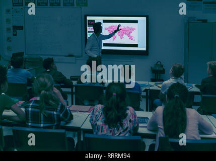 Gli studenti a guardare la geografia insegnante a schermo di proiezione in aula scuro Foto Stock