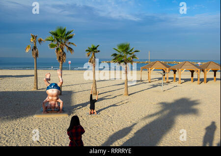 Giovane donna di scattare una foto del suo amico facendo un headstand accanto al Ben Gurion statua sulla spiaggia di Tel Aviv, Israele Foto Stock