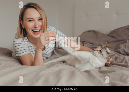 Ritratto di felice ridere caucasica sorridente donna bionda sdraiato sul letto in camera da letto a casa e giocare con la punta orientale-colorato cat. Proprietario di pet con Foto Stock