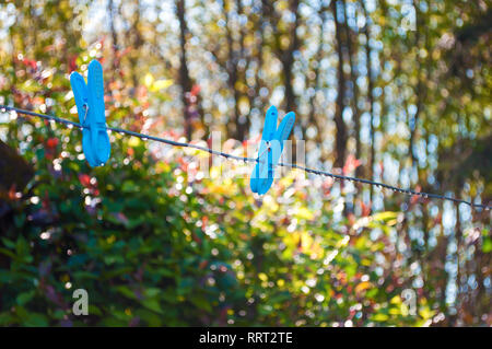 Blu di due pioli di vestiti appesi su una stringa di metallo con tante gocce di rugiada contro sfondo sfocato di un giardino e alberi ad alto fusto su una soleggiata mattina di primavera Foto Stock