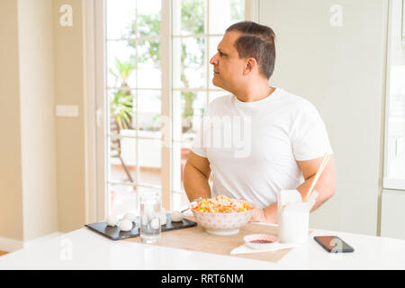 La mezza età uomo mangiare cibo asiatico con bacchette a casa cercando di lato, profilo relax posano con faccia naturale con sorriso sicuro. Foto Stock