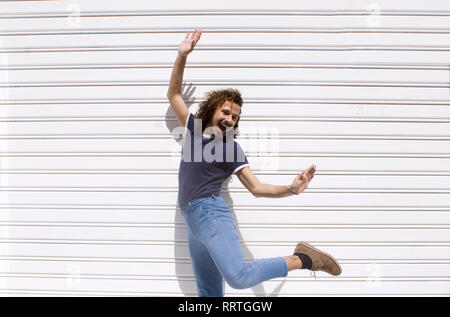 Allegra e felice giovane adulto con capelli ricci la barba e baffi a saltare e ballare in un muro bianco sullo sfondo. Elegante uomo gay Foto Stock