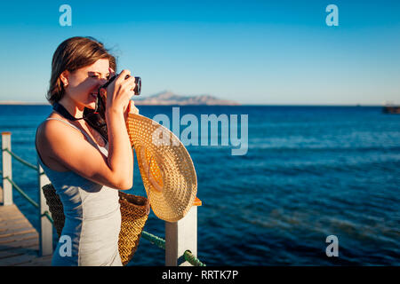 Giovane donna traveler per scattare delle foto del paesaggio di mare sul molo l'utilizzo della fotocamera. Moda estate. La vacanza estiva. Foto Stock