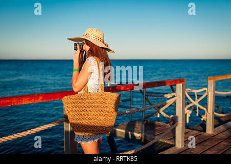 Giovane donna traveler per scattare delle foto del paesaggio di mare sul molo l'utilizzo della fotocamera. Moda estate. La vacanza estiva. Foto Stock