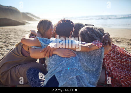 Abbracci di gruppo in cerchio sulla spiaggia soleggiata durante il ritiro di yoga Foto Stock