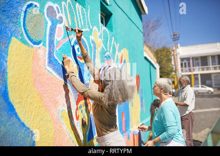 Senior volontari della comunità vibranti di pittura murale sulla soleggiata parete urbana Foto Stock