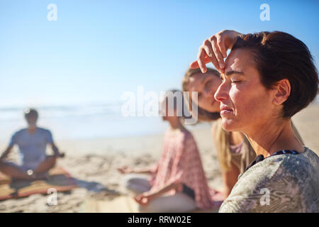 Istruttore Yoga toccando il terzo occhio di donna meditando sulla spiaggia soleggiata durante il ritiro di yoga Foto Stock