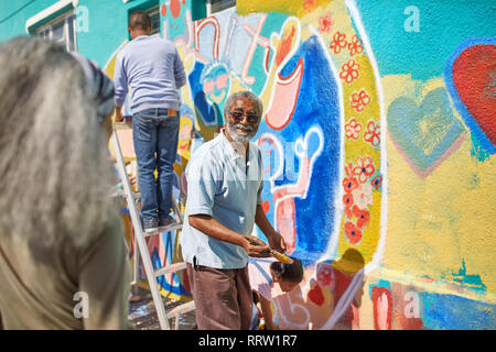 Senior uomo volontario vibrante di pittura murale sulla parete soleggiato Foto Stock