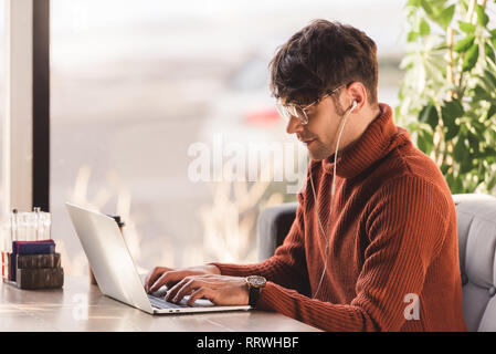 Felice giovane uomo che ascolta la musica in cuffia e utilizzando laptop in cafe Foto Stock