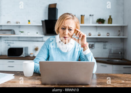 Scioccato senior donna tocca il viso, utilizzando laptop e avente la video chat in cucina Foto Stock