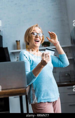 Sorpreso senior donna in bicchieri holding credito carta vicino al portatile a casa Foto Stock