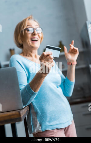 Excited donna senior in bicchieri holding credito carta vicino al portatile a casa Foto Stock