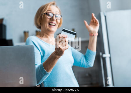 Excited donna senior in bicchieri gesti con le mani e la holding credito carta vicino al portatile a casa Foto Stock