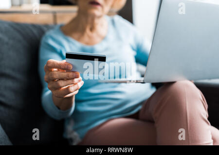 Vista ritagliata della senior donna seduta, tenendo la carta di credito e tramite computer portatile mentre facendo shopping online a casa Foto Stock