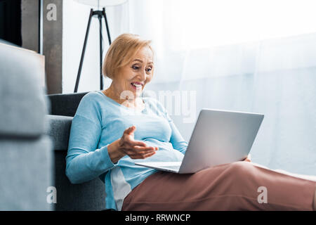 Sorpreso senior donna gesticolando con la mano mentre avente chiamata video su laptop a casa Foto Stock