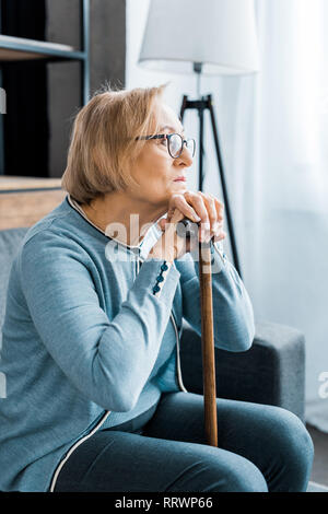 Premuto senior donna in bicchieri seduto con bastone e guardando lontano da casa Foto Stock