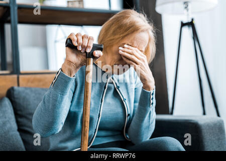 Sconvolto senior donna tenendo un bastone, che ricopre la faccia con la mano e pianto a casa Foto Stock