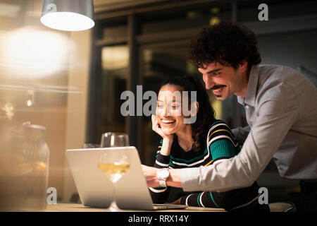Felice coppia utilizzando laptop e di bere il vino bianco a casa di notte Foto Stock