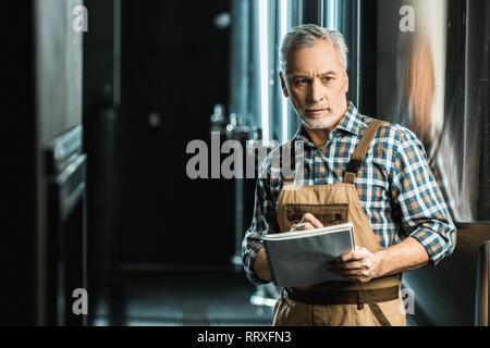 Professional senior brewer in tute da lavoro la scrittura nel blocco note nella fabbrica di birra Foto Stock