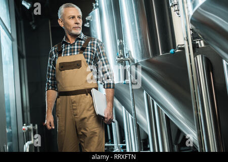 Birraio senior in tute da lavoro azienda notepad mentre esaminando la fabbrica di birra Foto Stock