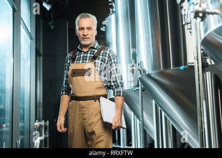 Professional senior brewer in tute da lavoro tenendo premuto il Blocco note e in posa nella fabbrica di birra Foto Stock