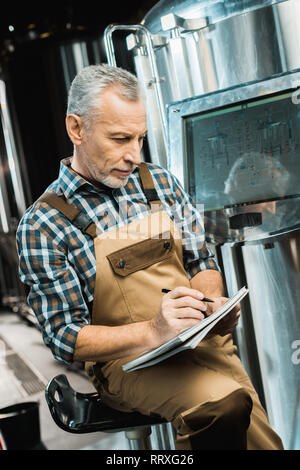 Birraio senior in tute da lavoro la scrittura nel blocco note mentre esaminando Birreria Attrezzature Foto Stock