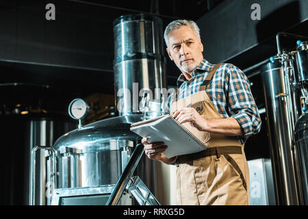 Birraio senior in tute da lavoro la scrittura nel blocco note mentre esaminando Birreria Attrezzature Foto Stock