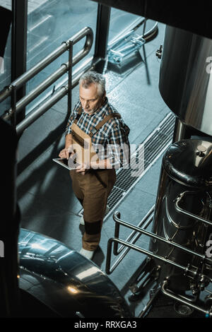 Vista aerea del birraio maschio in tute da lavoro con tavoletta digitale nella fabbrica di birra Foto Stock