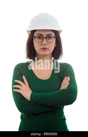 Gravi e fiducioso giovane donna ingegnere indossando occhiali e casco di protezione, tenendo le braccia incrociate isolate su sfondo bianco. Foto Stock