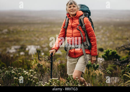 Avventurosa senior donna su un viaggio escursionistico. Senior donna in giacca e zaino trekking nella campagna tenendo un trekking pole. Foto Stock