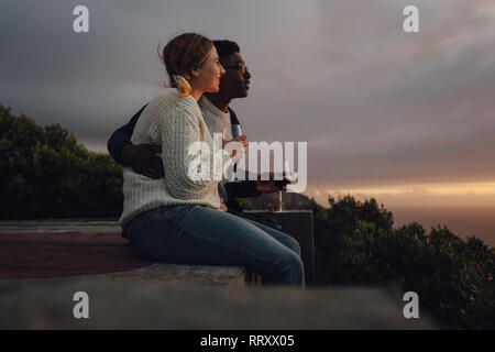 Vista laterale della romantica interracial giovane ammirando il tramonto. L uomo e la donna seduta insieme con bicchieri di vino. Foto Stock