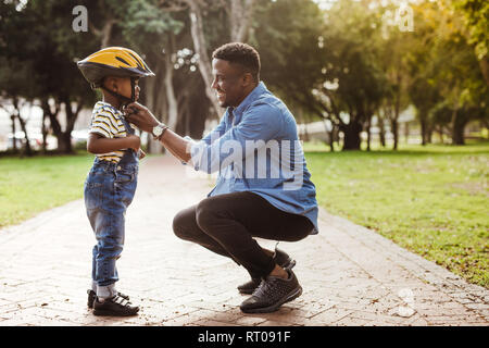 L'uomo africano di mettere il casco sulla cute boy al parco. Padre mette il figlio di un casco protettivo per l'equitazione Bicicletta. Foto Stock