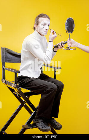 Attore di applicare il trucco per il viso per la pantomima sititng prestazioni in studio su sfondo giallo Foto Stock