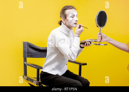 Attore di applicare il trucco per il viso per la pantomima sititng prestazioni in studio su sfondo giallo Foto Stock