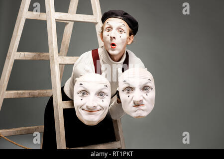 Ritratto di un attore pantoomime holding maschere facciali in studio con scaletta su sfondo grigio Foto Stock