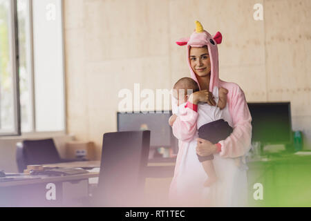 Giovane madre indossa unicorn onesie, stando in ufficio, tenendo il suo figlio nelle sue braccia Foto Stock