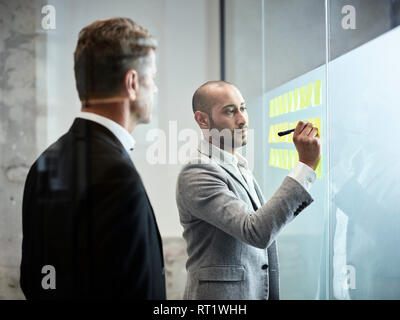 Imprenditore guardando collega iscritto su foglietti adesivi a lastre di vetro in ufficio Foto Stock