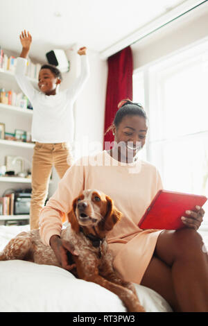 Giocoso ragazzo saltando sul letto dietro il cane e madre con tavoletta digitale Foto Stock