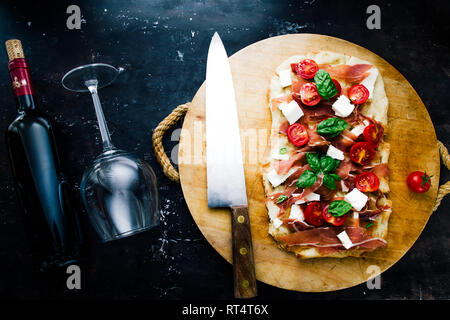 Artiginale pane piatto con pomodoro, Formaggio feta, prosciutto, e basilica, su un round tagliere su una scheda di colore nero, laici piatto con il coltello e il vino rosso Foto Stock