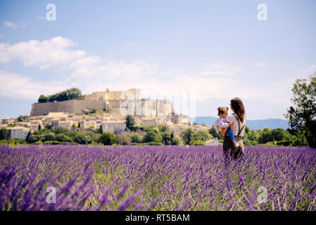 Francia, Grignan, madre in piedi nel campo di lavanda con piccola figlia sulle sue braccia guardando village Foto Stock