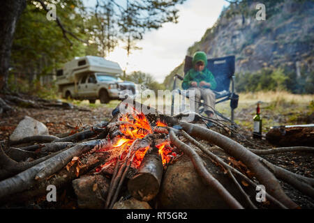 Il Cile, Santa Magda, Rio Maniguales, ragazzo seduto presso il fuoco nella foresta con camper in background Foto Stock