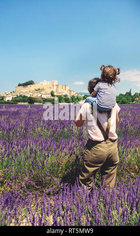 Francia, Grignan, vista posteriore della madre in piedi nel campo di lavanda con piccola figlia sulle sue spalle Foto Stock