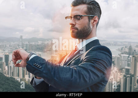 Il tempo di fare business. Bello barbuto imprenditore nel classico usura guardando il suo orologio mentre in piedi all'aperto con moderni edifici per uffici sullo sfondo. La gestione del tempo. Foto Stock