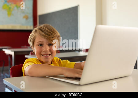 Felice ragazzo guardando la fotocamera durante l'utilizzo di laptop al banco in una classe Foto Stock