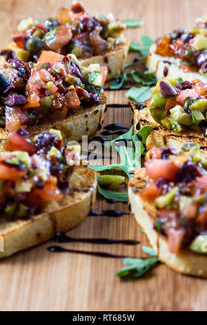 Bruschetta fresca fatta con una varietà di olive e cubetti di pomodoro su piccole fette biscottate di pane con una spruzzata di aceto balsamico riduzione su un vassoio in legno Foto Stock