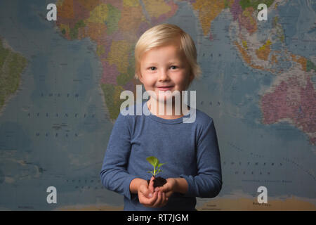 Carino biondo attentamente il bambino tiene una piantina di fronte ad una mappa del mondo Foto Stock