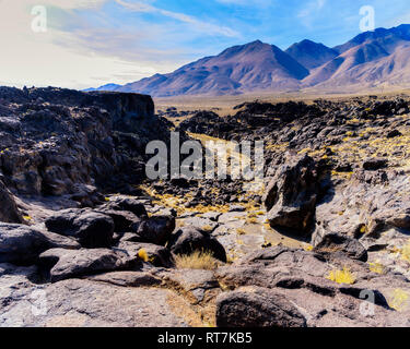 Cercando su terreno roccioso al roccioso canyon con frastagliate pareti di roccia che conduce al deserto arido valle e le montagne al di là sotto il luminoso cielo blu con nuvole bianche. Foto Stock