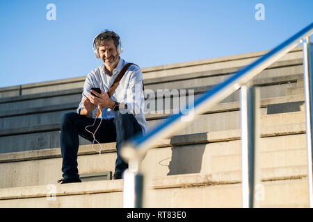 Ritratto di sorridere uomo maturo seduto sui gradini con le cuffie e il telefono cellulare Foto Stock