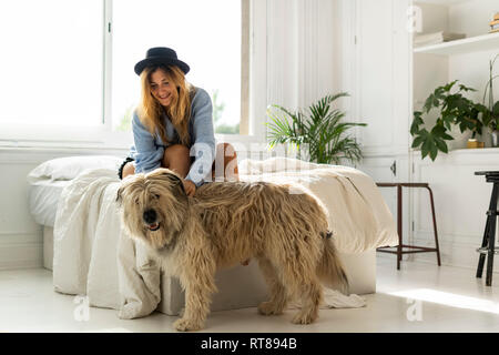 Sorridente giovane donna seduta sul letto di accarezzare un cane Foto Stock