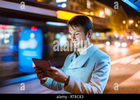 Imprenditore con tavoletta digitale sta in piedi in una fermata di autobus a notte Foto Stock